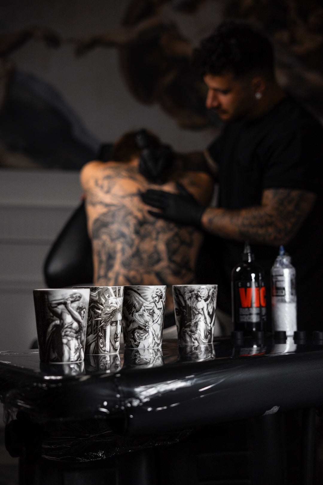 Andreas Ishak, tatuerare och designer av ICONCUPS-serien The Eternal Struggle, tatuerar en kunds rygg i sin studio medan muggar ur serien står uppställda på bordet i förgrunden.