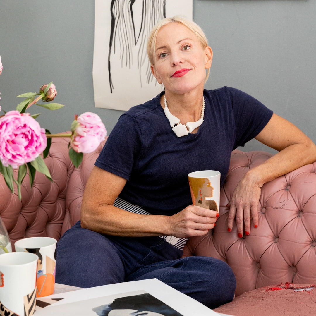 Stina Persson avslappnat sittande i sin studio, i rosa skinnsoffa med en ICONCUPS-mugg i handen – en kreativ ikon i sin rätta miljö.