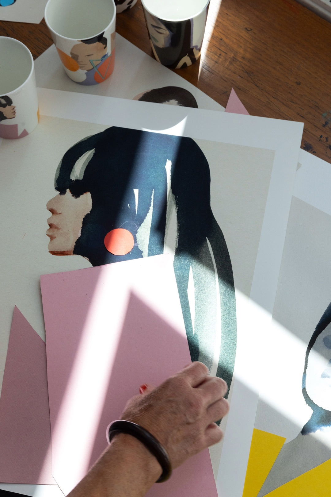 Stina Perssons hand i arbete i ateljén – visar processen bakom hennes konstverk, med färg, papper och skiss på arbetsbordet. ICONCUPS muggar skymtar i bakgrunden.