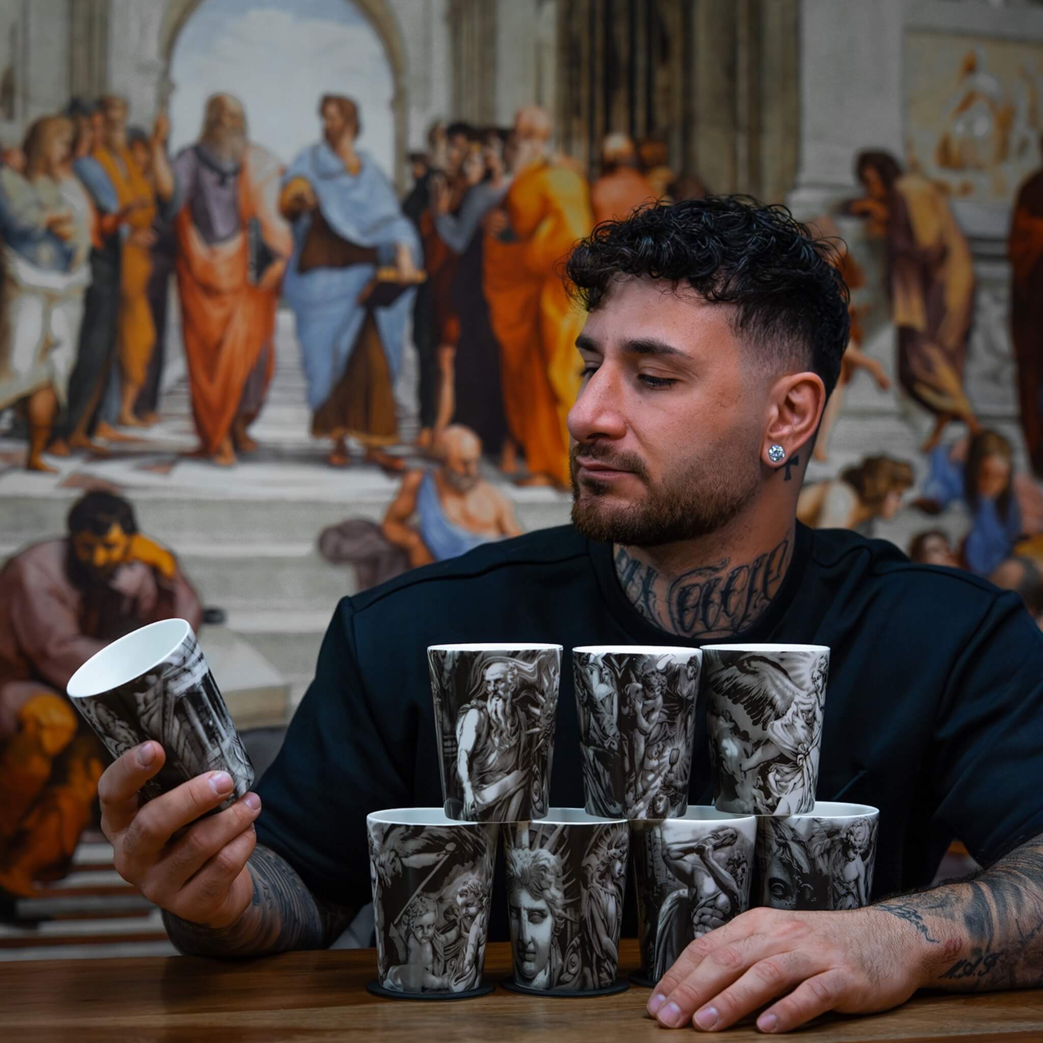 Andreas Ishak, tatuerare och designer av ICONCUPS-serien The Eternal Struggle, sitter vid ett bord med flera muggar ur serien och håller en av dem i handen medan han betraktar motivet.