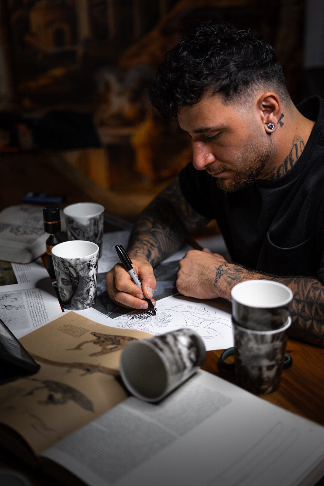 Andreas Ishak, tatuerare och designer av ICONCUPS-serien The Eternal Struggle, vid arbetsbord fyllt med inspirationsbilder, böcker och muggar ur serien medan han skissar fram motiven.