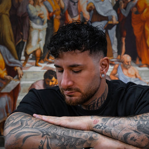 Tatueraren Andreas Ishak, designer av ICONCUPS-serien The Eternal Struggle, sittande med armarna i kors framför en tavla med figurer från antiken.