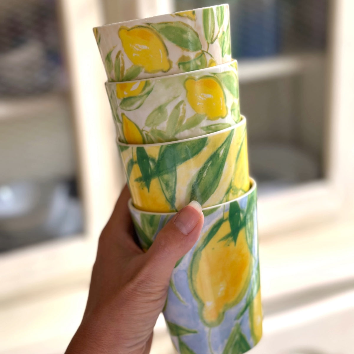 Muggar från serien ‘Citrons de Provence’ av Lena Linderholm – närbild där en hand håller muggar med vitrinskåp i bakgrunden